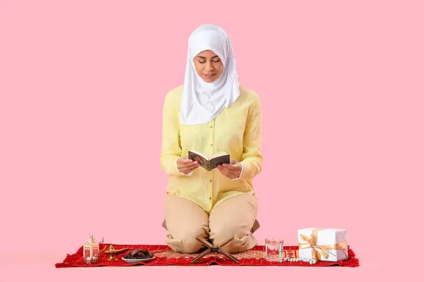Muslim woman sitting on prayer mat with Ramadan symbols and Koran on pink background