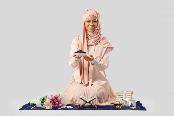 Muslim woman sitting on prayer mat with dried dates, Ramadan symbols and Koran on white background