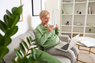 Evde kanepede oturan çay ve dizüstü bilgisayarı olan olgun bir kadın.