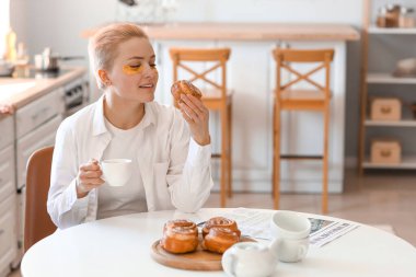 Bir fincan kahveyle mutfakta çörek yiyen genç bir kadın.