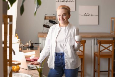 Mutfakta dizüstü bilgisayar kullanan bir fincan kahveli genç kadın.