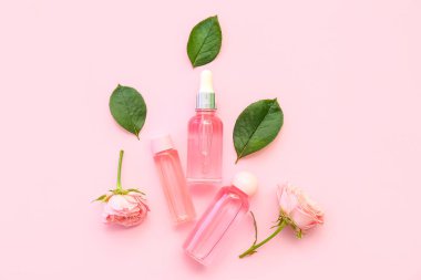 Gül özlü kozmetik yağı şişeleri ve pembe arka planda çiçekler.