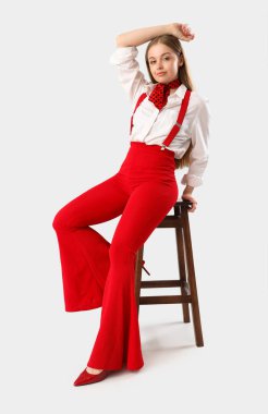 Askıları beyaz arka planda, taburede oturan şık genç bir kadın.