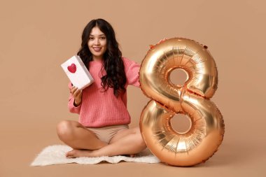Hediye kutusu ve kahverengi arka planda oturan 8 numaralı figür balonlu güzel, mutlu bir Asyalı kadın. Uluslararası Kadınlar Günü