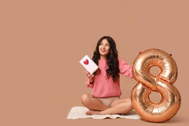Hediye kutusu ve kahverengi arka planda oturan 8 numaralı figür balonlu güzel, mutlu bir Asyalı kadın. Uluslararası Kadınlar Günü