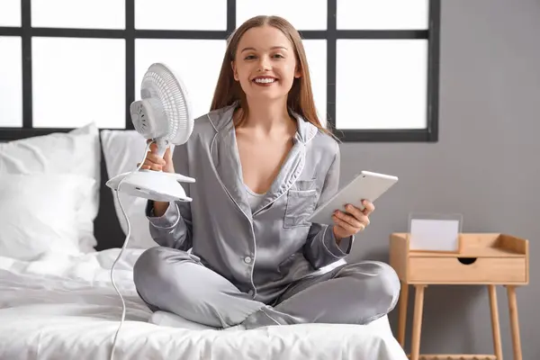 Young woman with tablet computer and electric fan in bedroom