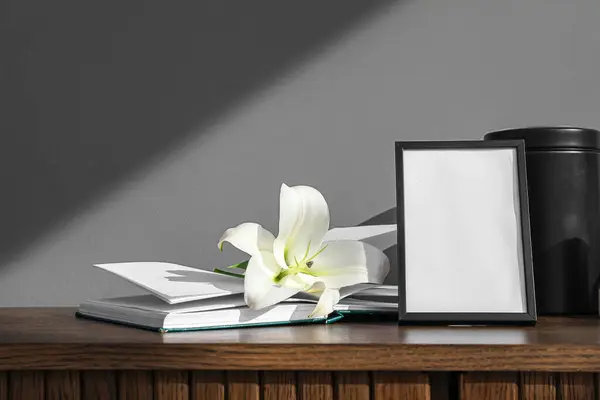 Blank funeral frame, book and lily flowers on wooden table near grey wall