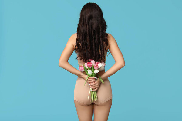 Beautiful young Asian woman in lingerie with bouquet of tulips on blue background, back view. International Women's Day