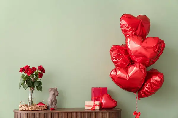 Wooden cabinet with bouquet of roses, gift boxes and heart-shaped balloons near green wall. Valentine\'s Day celebration