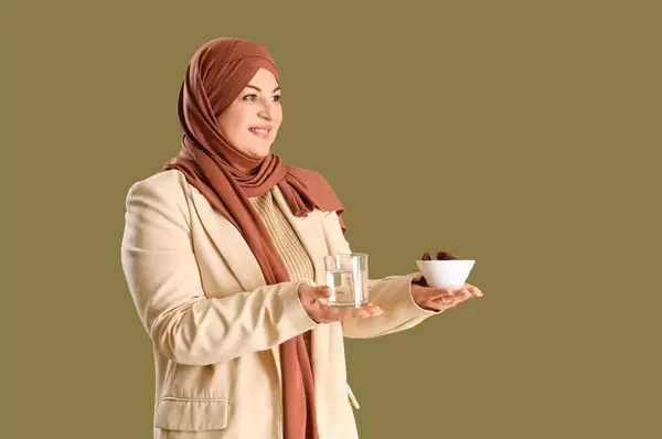 Mature Muslim woman with dates and glass of water on green background. Ramadan celebration