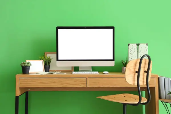 Modern workplace with blank computer monitor and artificial plants near green wall
