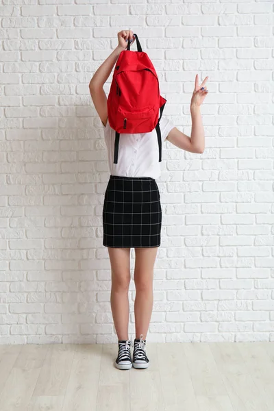 Female student with backpack showing victory gesture near white brick wall