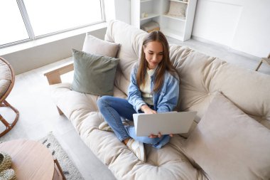 Oturma odasındaki koltukta dizüstü bilgisayar kullanan genç bir kadın.