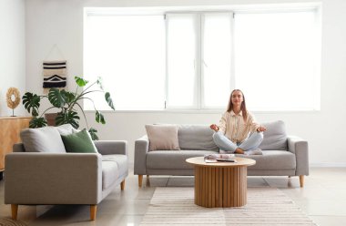 Genç bir kadın oturma odasında kanepede meditasyon yapıyor.