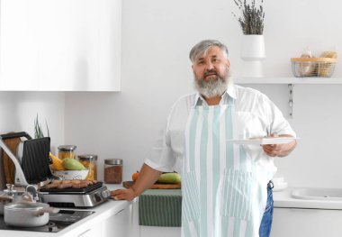 Mutfak masasındaki modern elektrikli ızgaranın üstünde pişmiş lezzetli sosislerle dolu tabağı olan olgun bir adam.