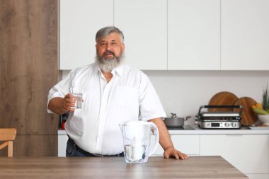 Mutfakta filtre sürahi ve bir bardak taze su olan olgun bir adam.