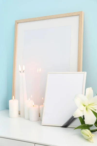 Blank funeral frame, beautiful lily flowers and burning candles on white cabinet near color wall, closeup