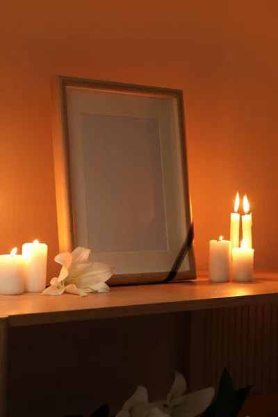Blank funeral frame, burning candles and lily flowers on wooden table in dark room