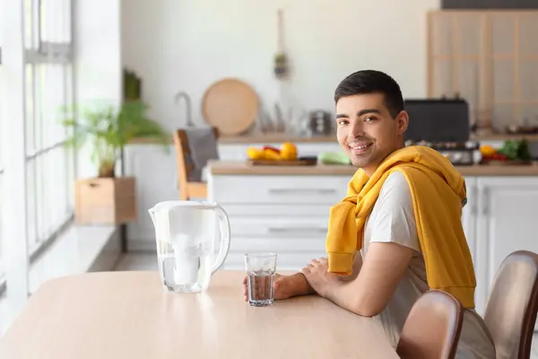 Young man with filter jug and glass of fresh water sitting at table in kitchen