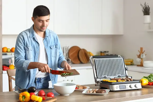 Young man cooking tasty sausages on modern electric grill and making salad of fresh vegetables in kitchen