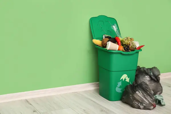 Container for trash with food waste and full garbage bags near green wall. Recycling concept