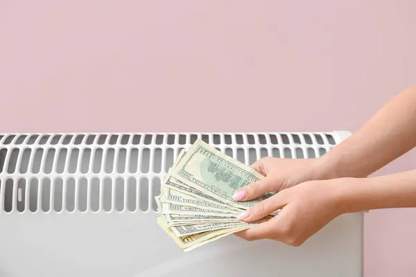 Female hands with money near electric convector heater near pink wall at home. Heating saving concept