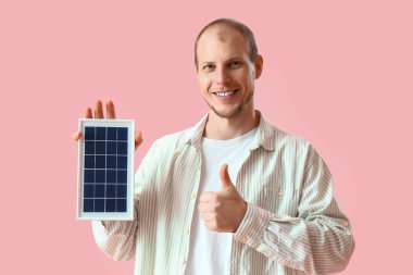 Portatif güneş paneli olan genç adam pembe arka planda baş parmağını kaldırıyor.