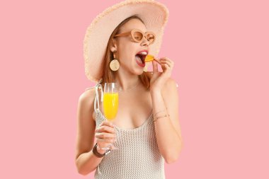 Hasır şapkalı güzel bir kadın bir bardak lezzetli narenciye kokteyli ile pembe arka planda portakal yiyor.
