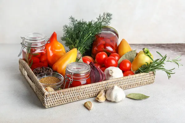 Wicker box with jars of pickled tomatoes and different spices on white background