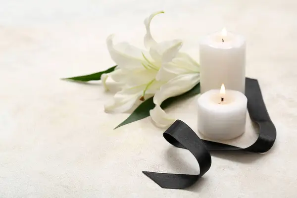 Burning candles, black funeral ribbon and lily flower on light background