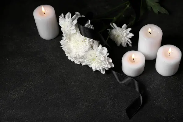 Burning candles, chrysanthemum flowers and black funeral ribbon on dark background