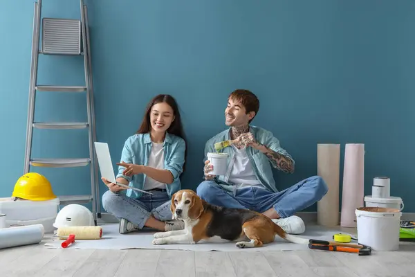 Young couple with painting tools and Beagle dog using laptop near blue wall during repair in their new house