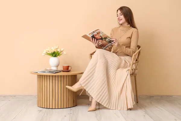 Beautiful woman reading fashion magazine in armchair near beige wall