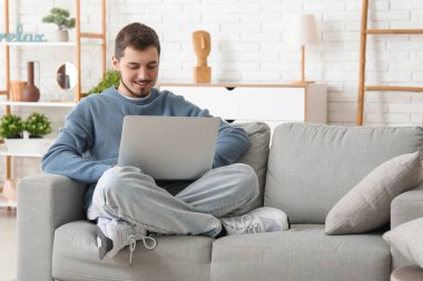 Oturma odasında kanepede otururken dizüstü bilgisayar kullanan yakışıklı genç adam.