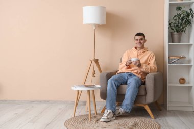 Elinde kahve fincanı olan genç adam evdeki koltukta oturuyor.