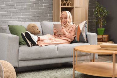 Küçük Müslüman kız evde kanepede yatarken tablet bilgisayar kullanıyor.
