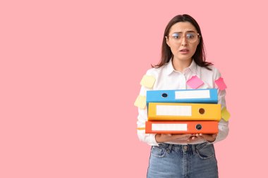 Pembe arka planda yapışkan notları ve belge klasörleri olan stresli genç iş kadını.