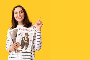 Dergileri olan genç bir kadın sarı arka plandaki bir şeyi işaret ediyor.