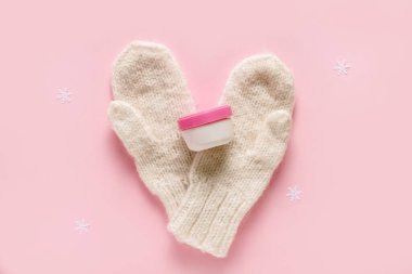 Kar taneli bir kavanoz krema ve pembe arka planda sıcak kış eldivenleri.