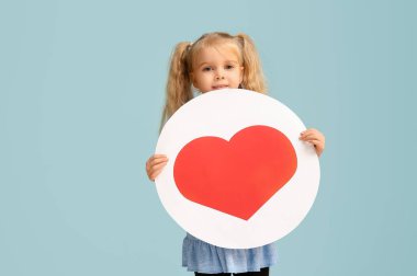 Mavi arka planda kırmızı kalbi olan küçük tatlı bir kız elinde büyük bir kart tutuyor. Sevgililer Günü kutlaması