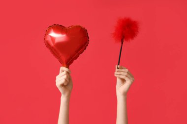 Kalp şeklinde balonu ve kırmızı arka planda seks dükkanından tüy çubuğu olan kadın elleri. Sevgililer Günü kutlaması