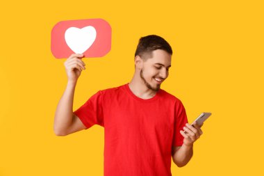Sarı arka planda ikon ve cep telefonu gibi kalbi olan yakışıklı genç bir adam.
