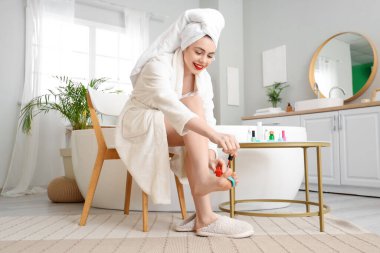 Güzel, genç ve mutlu bir kadın, elinde bir şişe ojeyle banyoda pedikür yapıyor.