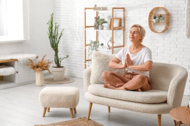 Olgun bir kadın evde kanepede otururken meditasyon yapıyor.