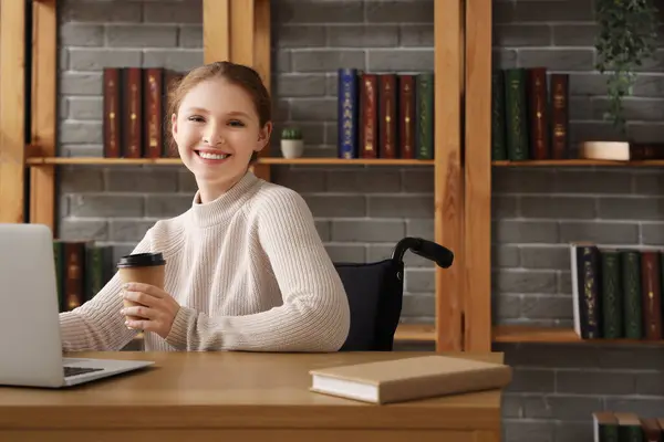 Young redhead woman in wheelchair with coffee cup using laptop at library