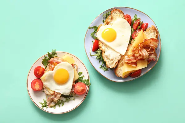 Plates with tasty fried eggs, toasts, tomatoes and arugula on turquoise background