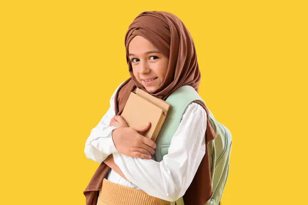 Little Muslim girl in hijab with school backpack and books on yellow background