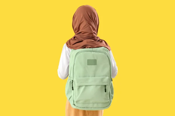 Little Muslim girl in hijab with school backpack on yellow background, back view