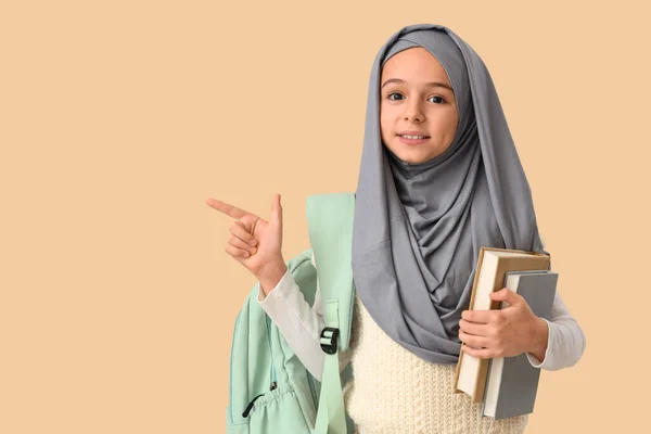 Little Muslim girl in hijab with school backpack and books pointing at something on beige background