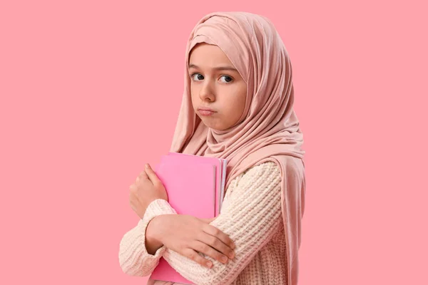 Sad Muslim girl in hijab holding copybooks on pink background